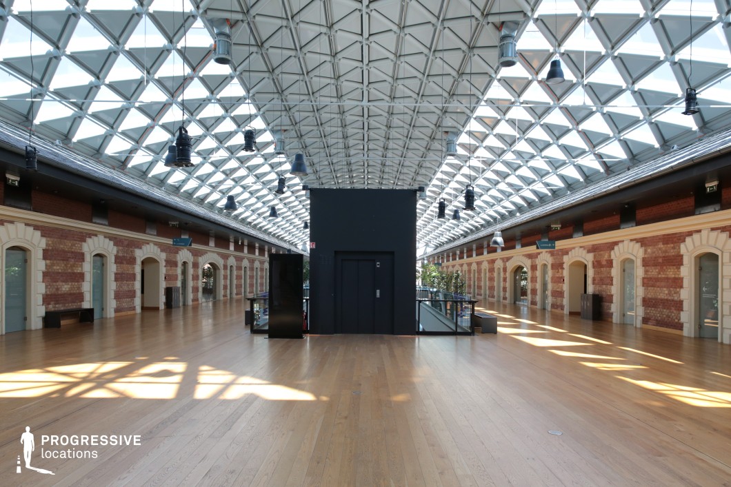 This expansive atrium seamlessly blends classical architecture with modern industrial elements. The symmetrical space is characterized by a vaulted glass ceiling composed of a geometric metal framework, allowing natural light to flood the open area. The walls feature exposed red brick with elegant white stone detailing around arched doorways, creating a contrast between heritage and contemporary design. The wooden flooring adds warmth, while the centrally located black elevator shaft serves as a stark modern centerpiece. Industrial-style hanging lights emphasize the venue's hybrid aesthetic. This location is well-suited for high-end fashion shoots, futuristic narratives, or corporate thrillers, providing an adaptable backdrop for dramatic entrances, tense confrontations, or elegant gatherings.