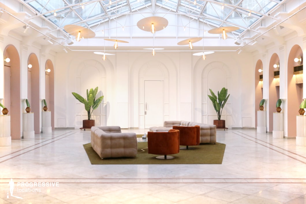 This refined lounge space features a high glass ceiling supported by white steel framework, allowing an abundance of natural light to illuminate the room. The architecture blends neoclassical elements with modern touches, including arched alcoves, decorative wall paneling, and soft pastel accents. The polished marble floor is contrasted by a central seating arrangement consisting of plush beige sofas and burnt-orange swivel chairs on a muted green rug. Large potted plants enhance the organic feel of the space, while sculptural pendant lights add warmth and sophistication. This location is ideal for luxury lifestyle shoots, high-end fashion campaigns, or dramatic film scenes that require an opulent yet contemporary setting, perfect for elite gatherings, intimate conversations, or artistic expositions.