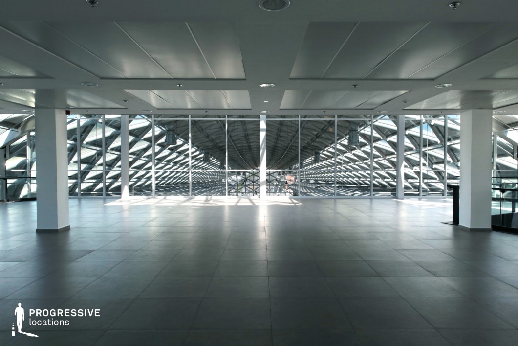 This expansive, modern space features a high-tech glass and steel structure with a geometric, tunnel-like ceiling that extends into the distance. The open floor is covered in large, neutral gray tiles, while the clean white ceiling incorporates recessed lighting for a sleek, minimalist look. The vast glass facade allows natural light to cast dynamic shadows across the floor, creating a futuristic and high-contrast atmosphere. The rigid symmetry and industrial materials make this an ideal setting for sci-fi productions, corporate thrillers, or dystopian narratives. Its sterile, high-tech aesthetic is well-suited for high-stakes negotiations, surveillance sequences, or dramatic chase scenes in a modern or futuristic world.
