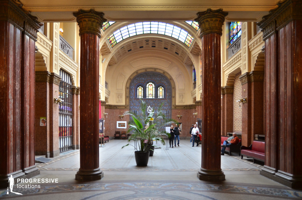 This grand hall blends classical and Art Nouveau architectural elements, featuring towering polished marble columns with ornate capitals that frame the space. The arched ceiling is accentuated by a stained-glass skylight, allowing natural light to illuminate the warm-hued stone walls and decorative mosaics. The focal point at the far end is a large ornamental wall with intricate blue and gold tilework, complemented by arched stained-glass windows that add vibrancy to the setting. The symmetrical layout, refined materials, and soft ambient lighting create an elegant yet welcoming atmosphere. This location is ideal for filming historical dramas, museum or cultural settings, sophisticated social gatherings, or artistic and intellectual discussions, offering a visually rich and culturally significant backdrop.