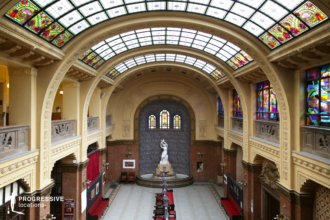 This magnificent Art Nouveau hall features a vast arched ceiling adorned with intricate stained glass panels that filter natural light into the space. The symmetrical design is enhanced by decorative columns, detailed balustrades, and warm-toned walls with ornamental moldings. At the far end, a central alcove showcases a classical marble statue set against a richly tiled mosaic backdrop, framed by three arched stained-glass windows. The polished stone flooring, red velvet seating, and exhibition-style displays contribute to the hall’s refined yet historic character. This location is ideal for filming historical dramas, period thrillers, museum-based narratives, or elegant formal gatherings, providing a visually rich and atmospheric setting.
