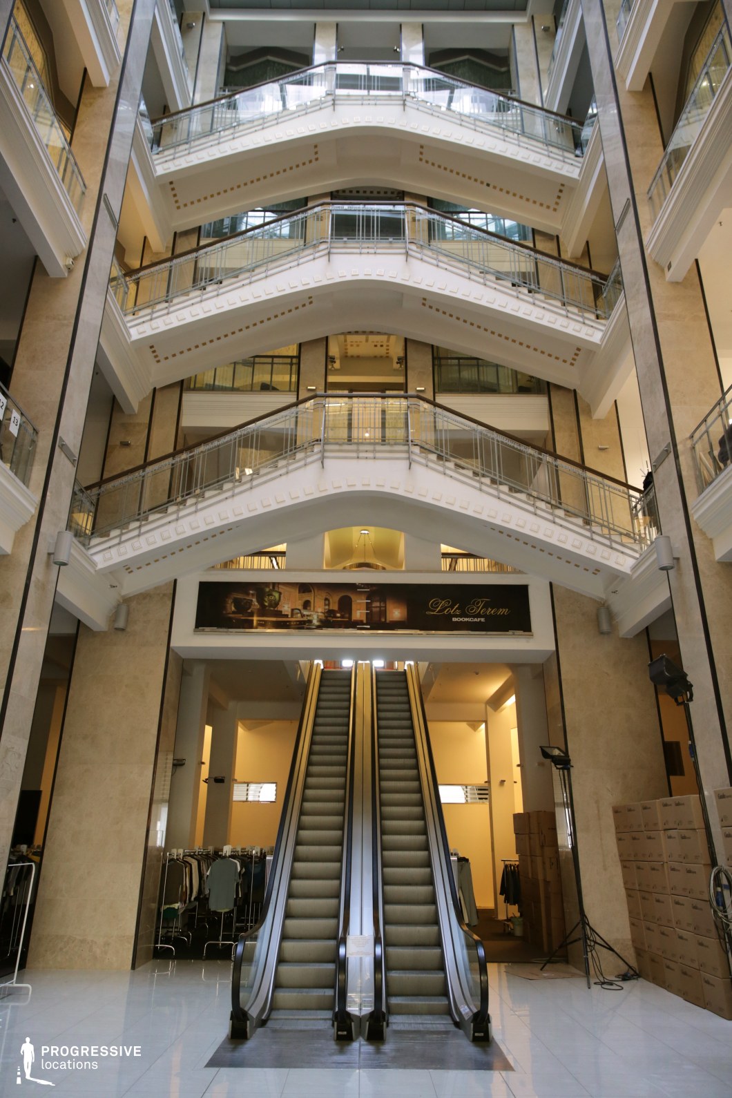 This striking atrium features a pair of centrally positioned escalators leading up to multiple levels of curved balconies with elegant metal railings. The symmetrical design, accentuated by beige marble walls and polished white flooring, exudes a refined yet functional aesthetic. The high ceilings and layered architecture create a sense of openness, while the art deco-inspired details on the balconies add a touch of classic grandeur. Retail elements and a book café sign hint at a mixed-use commercial space. This location is well-suited for filming corporate dramas, high-society intrigue, or action sequences involving chases and suspenseful encounters in a grand yet neutral urban setting.
