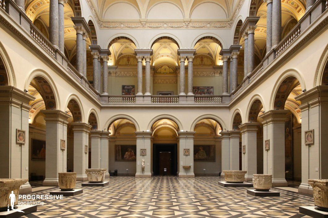 This stately museum hall embodies neoclassical elegance, featuring towering marble columns, arched openings, and a symmetrical two-tiered gallery design. The intricate geometric-patterned marble flooring enhances the space’s grandeur, while the vaulted ceilings adorned with ornamental frescoes add an air of refinement. Soft, ambient lighting highlights the architectural details and classical artwork displayed along the walls. The open layout, with its rhythmic repetition of columns and arches, creates a visually striking depth and balance. This location is ideal for filming historical dramas, art heist thrillers, and period mysteries, making it a perfect setting for scenes involving grand unveilings, intellectual debates, or high-society intrigue.