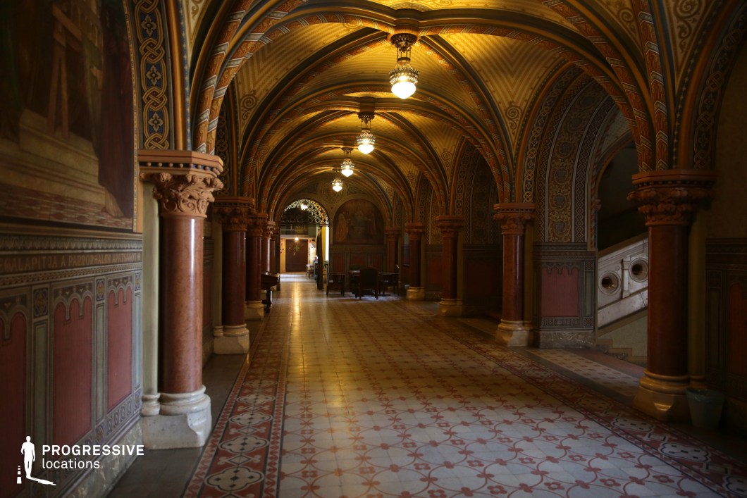  This stunning corridor features a series of elegant ribbed vaults supported by intricately carved columns with richly detailed capitals. The walls and ceilings are adorned with elaborate geometric patterns, evoking a Neo-Romanesque or Gothic Revival aesthetic. Decorative tile flooring with intricate motifs enhances the historic ambiance, while arched entryways create depth and perspective. Hanging lantern-style chandeliers cast a warm, atmospheric glow, accentuating the rich color palette of deep reds, golds, and earth tones. This location is ideal for filming historical dramas, mystery thrillers, or scenes set in a grand palace, monastery, or secretive medieval archive.