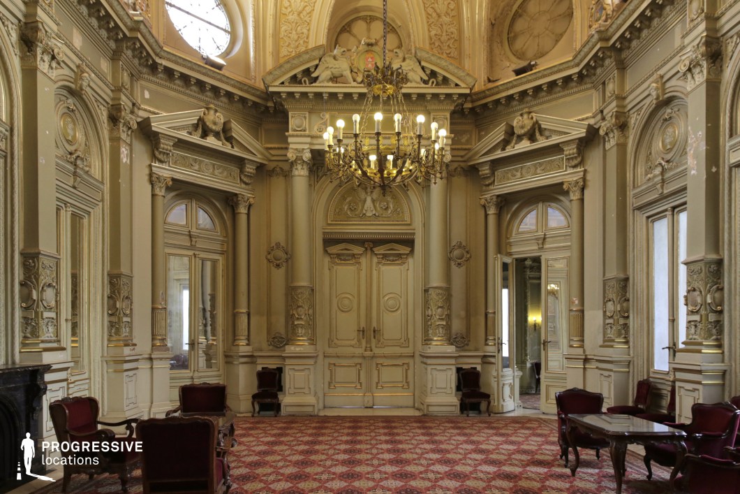 This lavish neoclassical salon is adorned with intricate architectural details, featuring grand double doors framed by Corinthian columns, gilded moldings, and sculptural reliefs that exude aristocratic elegance. The high, domed ceiling is crowned by a grand chandelier, casting a warm glow over the room. Large arched mirrors enhance the sense of space, reflecting the light and emphasizing the ornate plasterwork. The walls, painted in muted gold and cream tones, are complemented by deep red patterned carpeting and dark wood antique furniture. This location is ideal for filming historical dramas, royal court scenes, or aristocratic intrigue, offering a refined setting for diplomatic meetings, secret rendezvous, or high-society gatherings.