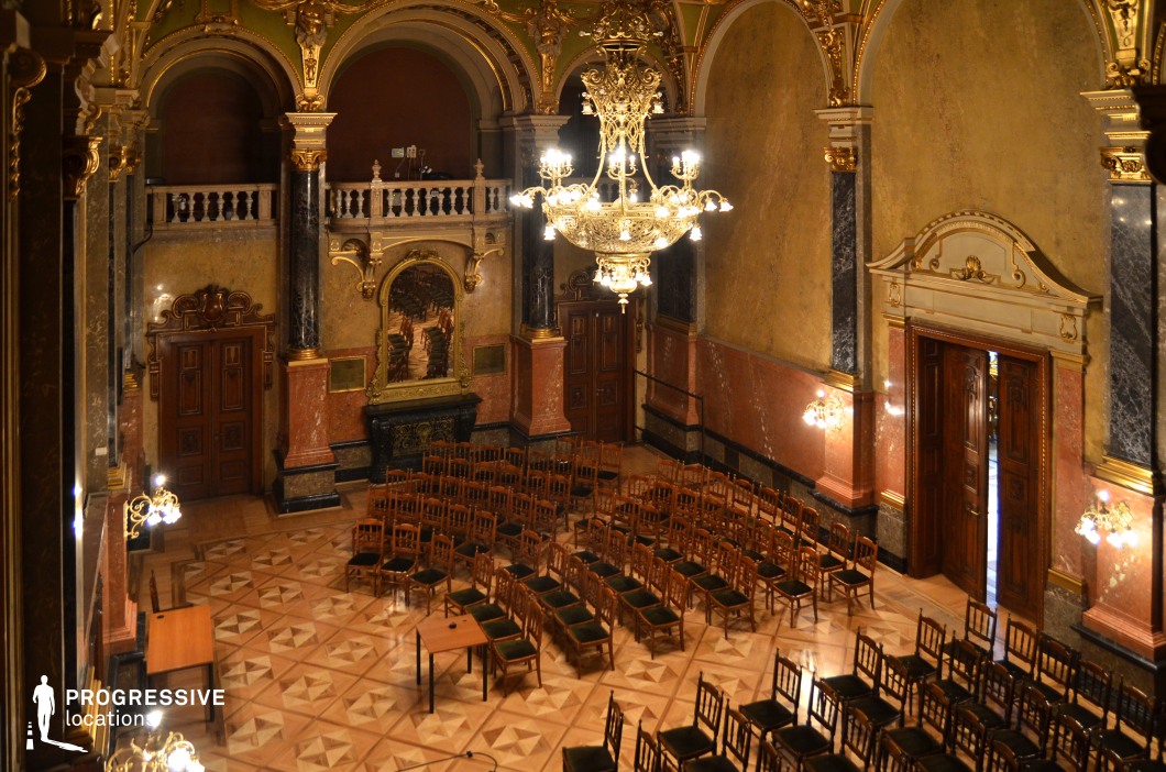This grand hall showcases an exquisite blend of Baroque and Rococo architectural elements, featuring soaring arched ceilings adorned with gold embellishments and intricate detailing. The marble-clad walls, rich in warm earth tones, contrast elegantly with the polished parquet flooring, which displays an intricate geometric pattern. The focal point is the opulent gilded chandelier, radiating a warm glow over the rows of wooden chairs arranged for formal gatherings. Large wooden double doors with elaborate carvings, along with arched balconies and decorative moldings, add to the regal atmosphere. This location is ideal for filming period dramas, political or royal court scenes, and high-society gatherings, providing an authentic and visually captivating setting.