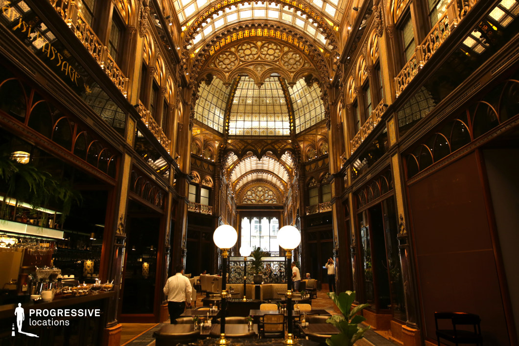 This breathtaking arcade showcases intricate neo-Gothic and Art Nouveau elements, with gilded detailing, arched windows, and an opulent glass ceiling that floods the space with natural light. The symmetrical design emphasizes grandeur, featuring elaborate ironwork, decorative motifs, and a series of repeating arches leading towards a luminous focal point. The polished wooden surfaces, reflective mirrors, and ambient lighting create a warm yet sophisticated atmosphere. The space, housing a refined café and lounge area, blends historic elegance with modern luxury. This location is ideal for filming high-society gatherings, period dramas, noir-style thrillers, or lavish historical narratives, offering an exquisite setting that evokes timeless prestige.