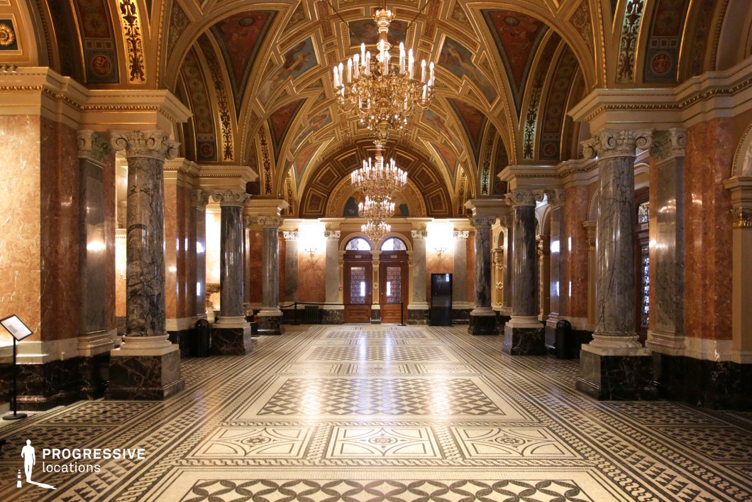 This opulent hall is defined by its soaring vaulted ceilings adorned with intricate frescoes, gold detailing, and symmetrical chandeliers that illuminate the space. The polished marble columns, in varying hues of deep gray and warm beige, add a sense of grandeur, while the geometric-patterned mosaic flooring enhances the regal aesthetic. The double wooden doors framed by arched windows serve as an elegant focal point, further emphasizing the hall’s classical architectural influence. The combination of rich textures, refined symmetry, and historical opulence makes this location ideal for filming period dramas, royal court scenes, high-society gatherings, or fantasy epics requiring a majestic and aristocratic setting.