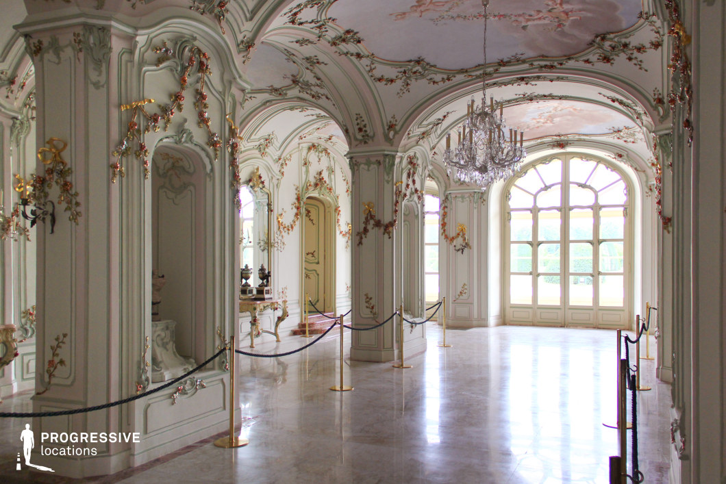 This grand Rococo-style hallway features delicate pastel-hued walls adorned with intricate floral gilded moldings and sculpted details. The high, arched ceilings are embellished with soft frescoes, while a crystal chandelier adds an air of refinement. Large, arched windows framed by elegant curved paneling allow natural light to flood the space, highlighting the polished marble flooring. Ornate mirrors, decorative consoles, and gold accents contribute to the luxurious ambiance. The space's open layout, flowing architecture, and artistic detailing make it an ideal filming location for historical dramas, period pieces, fantasy productions, and scenes requiring a regal or aristocratic atmosphere.