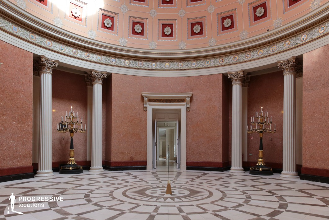 This stately neoclassical rotunda features a domed ceiling adorned with intricate floral medallions and ornamental friezes, set against warm red and peach hues. Tall, fluted Corinthian columns encircle the space, complementing the polished marble walls and a geometric-patterned marble floor with a central focal point. Two grand candelabras with gilded detailing enhance the sense of elegance, while an open doorway framed by decorative molding offers a glimpse into adjoining rooms, adding depth to the composition. This location is ideal for filming historical dramas, period epics, or regal ceremonies, providing a sophisticated setting for political discourse, aristocratic encounters, or philosophical debates.