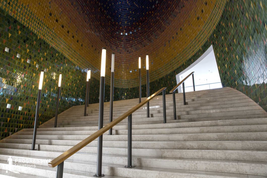 This striking staircase is enveloped by curved walls covered in a vibrant mosaic of small, triangular tiles in shades of green, gold, and deep blue, creating a rich, immersive environment. The wide stone steps ascend toward a bright, open space, flanked by sleek metal railings with wooden handrails. Tall, cylindrical light fixtures with a minimalist design punctuate the steps, adding a futuristic ambiance. The combination of intricate mosaic work, geometric precision, and soft illumination makes this location ideal for filming elegant, dramatic, or surreal sequences, fitting for science fiction, fantasy, or high-art cinematography.