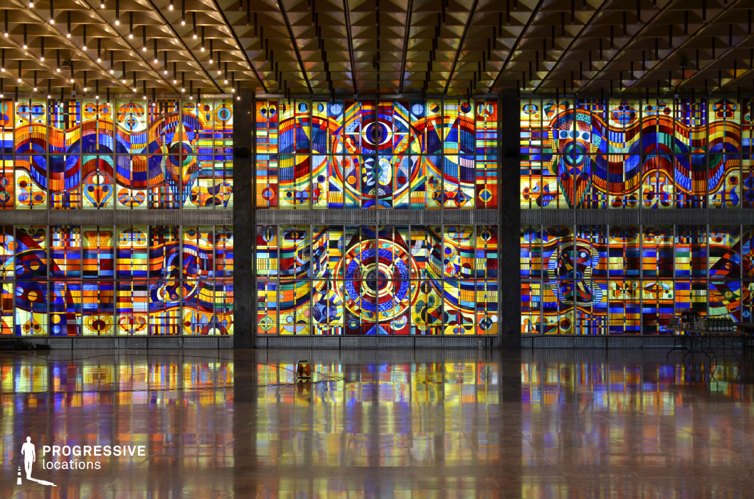 This expansive hall is dominated by an enormous stained-glass installation that stretches across the entire wall, featuring vibrant geometric patterns, abstract shapes, and a mesmerizing interplay of colors. The glass panels create a kaleidoscopic effect as natural and artificial light filter through, casting intricate reflections onto the highly polished marble floor. The coffered ceiling, adorned with a repetitive geometric pattern and embedded lighting, adds to the structured yet artistic ambiance. Dark vertical columns provide contrast, grounding the space in a bold architectural rhythm. This visually striking location is ideal for filming grand, surreal, or visionary sequences, including sci-fi, avant-garde, or high-art productions. It is well-suited for scenes depicting futuristic institutions, ceremonial spaces, or dreamlike encounters.