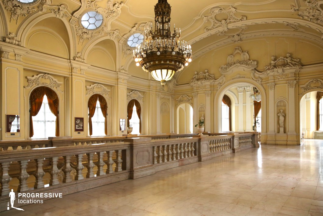 This elegant baroque-style hallway is adorned with intricate stucco moldings, sculptural reliefs, and arched openings that create a sense of grandeur and fluidity. The high, curved ceiling features decorative circular windows that allow soft, diffused light to enhance the warm yellow tones of the walls. A large, ornate chandelier hangs at the center, illuminating the polished marble flooring and intricately carved balustrades. Rich drapery in deep gold accents the tall windows, adding to the regal ambiance. This location is ideal for filming scenes requiring a majestic palace setting, making it perfect for historical dramas, aristocratic intrigue, or period-piece dance sequences.