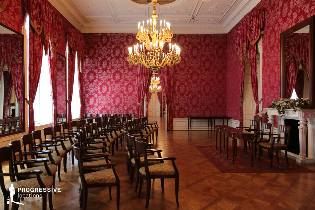 This luxurious hall exudes aristocratic elegance with its rich red damask-patterned walls, heavy drapery, and grand chandeliers casting a warm glow over the space. The parquet flooring enhances the room’s refined atmosphere, while a stately white marble fireplace adorned with decorative floral arrangements adds a touch of sophistication. Rows of classic wooden chairs suggest a formal gathering or event setting, with an arrangement that draws attention toward the front of the room. Large mirrors framed in dark wood amplify the grandeur, reflecting the intricate details of the interior. This location is ideal for filming historical dramas, royal meetings, political intrigue, or high-society gatherings, offering an opulent yet intimate ambiance.