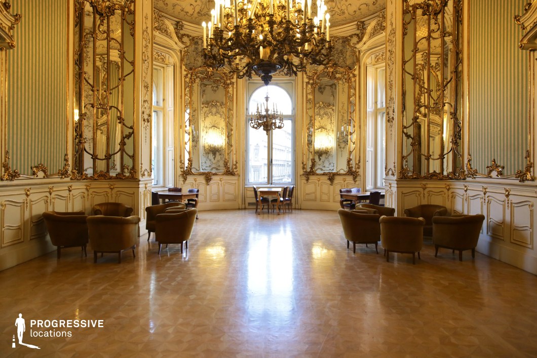 This grand ballroom exudes opulence with its intricate Baroque-style decor, characterized by gilded moldings, elaborate wall mirrors, and high, arched windows that allow natural light to accentuate the rich detailing. The ceiling hosts an elaborate chandelier, casting a warm glow over the polished parquet flooring. The soft pastel green and gold color palette enhances the regal atmosphere, while plush velvet seating areas create an inviting yet formal ambiance. The symmetry of the room, combined with the reflective surfaces, enhances its sense of depth and grandeur. This location is ideal for filming historical dramas, period pieces, or high-society gatherings, making it a perfect backdrop for scenes of aristocratic life, refined elegance, or secret rendezvous in a lavish setting.