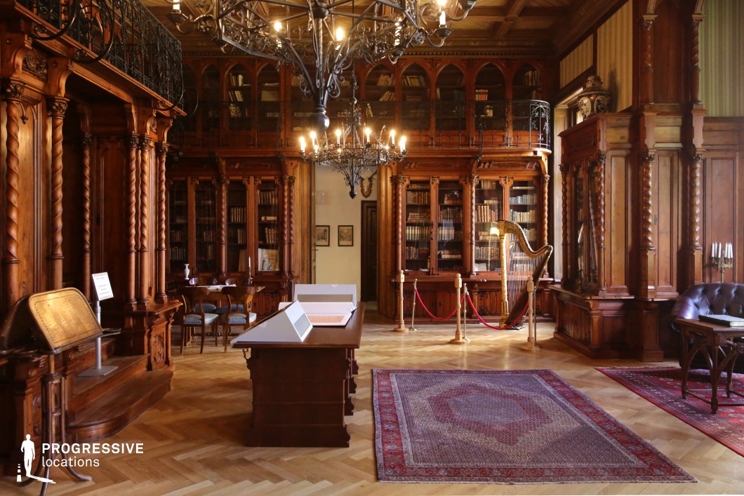 This luxurious historic library exudes old-world elegance, featuring intricately carved wooden bookshelves that rise to a mezzanine level with wrought-iron railings. The warm-toned wood is complemented by detailed columns, arched glass cabinet doors, and a coffered ceiling adorned with decorative carvings. Chandeliers cast a soft glow, illuminating the rich parquet flooring, antique rugs, and classical furnishings. A harp displayed in the corner adds to the refined atmosphere, enhancing the scholarly ambiance. This location is ideal for filming historical dramas, aristocratic intellectual gatherings, or scenes of intrigue set in an opulent private study or grand estate library.