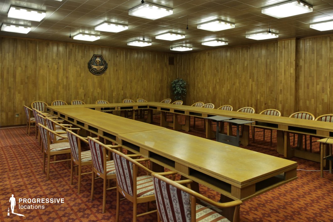 This mid-to-late 20th-century conference room features wood-paneled walls and a distinctive geometric ceiling lined with square recessed lighting. The space is dominated by a large U-shaped wooden table surrounded by vintage-style chairs with wooden frames and patterned upholstered seats. The deep red patterned carpet adds to the warm, retro aesthetic, reinforcing the setting’s institutional or corporate ambiance. A decorative emblem on the wall serves as a focal point, suggesting an official or governmental function. The symmetrical arrangement of furniture and lighting provides a formal yet nostalgic atmosphere, making this location ideal for filming historical dramas, political thrillers, or corporate period pieces. The setting is well-suited for scenes depicting board meetings, secret negotiations, or retro-themed office environments.