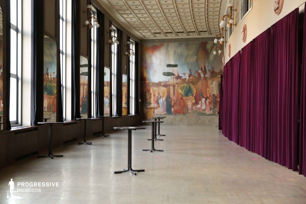 This spacious, high-ceilinged hall features a sweeping curved wall lined with large windows, allowing natural light to accentuate the elegant interior. A grand fresco dominates the far end, depicting a historical scene with warm, earthy tones, reinforcing the hall's classical aesthetic. The ceiling showcases a grid of recessed square panels, contributing to a structured and stately atmosphere. Ornate wall-mounted light fixtures add a vintage touch, while the deep red stage curtains introduce a theatrical element. This location is ideal for period dramas, historical reconstructions, or sophisticated social gatherings in film, lending itself well to formal receptions, diplomatic events, or artistic discussions.