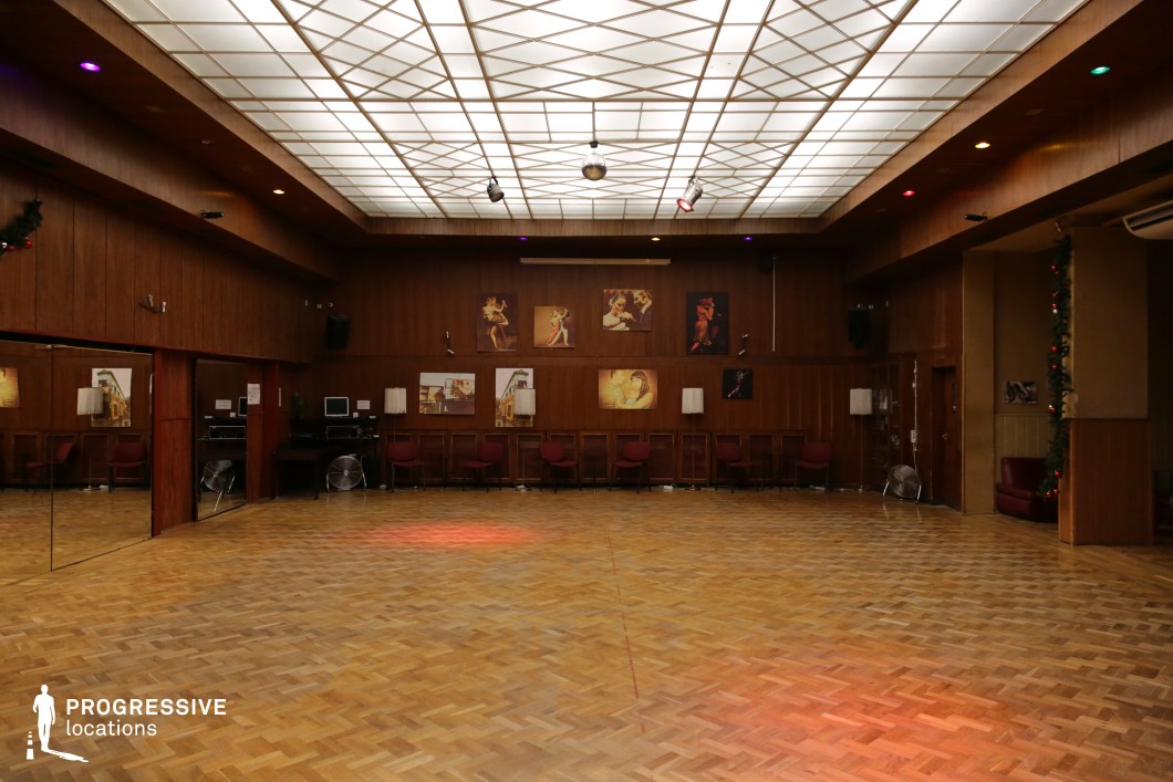 This spacious dance hall exudes a mid-century charm with its wood-paneled walls, parquet flooring, and large mirrored surfaces that enhance the sense of depth. A geometric-patterned illuminated ceiling provides soft, ambient lighting, while subtle colored spotlights add a dynamic touch. The room is adorned with vintage-style artwork depicting dancers, reinforcing its function as a social or performance space. A row of seating along the walls, a small DJ or sound booth, and festive garlands contribute to its nostalgic character. This location is ideal for filming retro-themed dance sequences, social gatherings, music videos, or period dramas set in the late 20th century, offering an authentic and inviting backdrop.