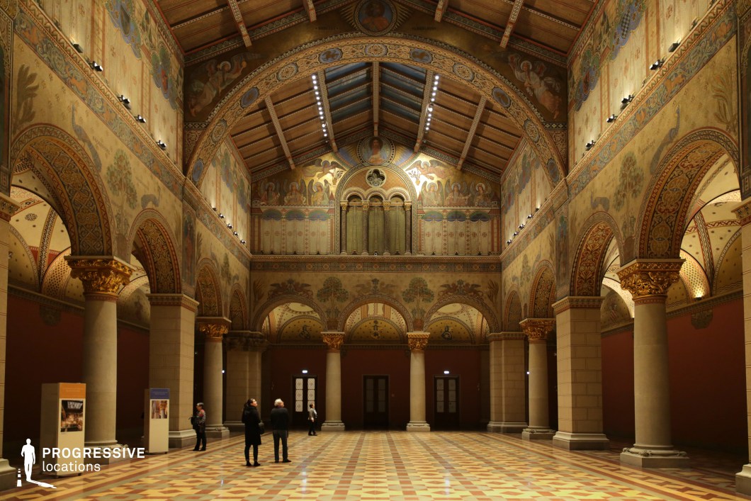 This grand hall features high vaulted ceilings adorned with intricate frescoes, decorative arches, and richly detailed patterns inspired by Romanesque and Byzantine art. The walls are embellished with ornate, nature-inspired motifs and religious iconography, while towering stone columns with gilded capitals provide a sense of grandeur and historical authenticity. The polished checkered tile flooring reflects the warm ambient lighting, enhancing the hall’s regal atmosphere. The symmetrical layout and rhythmic repetition of arches create a visually striking perspective. This location is ideal for filming historical dramas, period pieces, and fantasy productions. It can serve as a setting for royal court scenes, sacred ceremonies, or mysterious, esoteric gatherings.