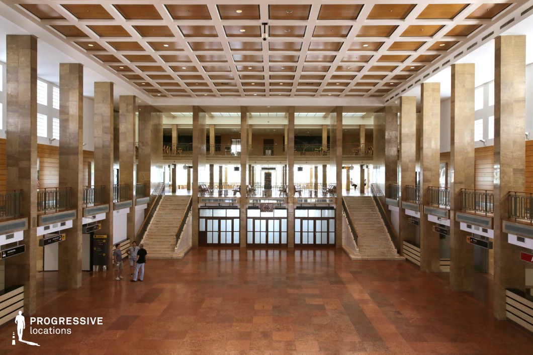 This imposing hall features a striking mid-century modernist design, characterized by towering marble-clad columns, a symmetrical layout, and a high coffered ceiling with wooden paneling. The polished stone floors and wide staircases leading to an upper level create a sense of grandeur and authority. Large frosted-glass doors at the far end, along with high-set windows, allow natural light to illuminate the expansive space. The clean lines, rigid symmetry, and functionalist design evoke a historical institutional setting. This location is ideal for filming political thrillers, Cold War-era dramas, dystopian settings, or high-stakes espionage sequences, offering a vast and formal yet slightly austere atmosphere.