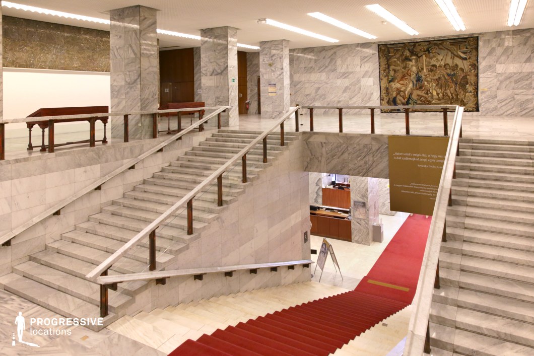 This grand interior features a striking multi-tiered marble staircase, enhanced by a dramatic red carpet that guides the eye downward to a lower-level reception area. The walls and floors are clad in polished white and gray marble, contributing to an austere yet prestigious ambiance. A large woven tapestry depicting a historical scene adds a rare decorative element, contrasting with the rigid geometric forms of the architecture. The space is illuminated by recessed linear lighting, reinforcing the structured, institutional aesthetic. This location is ideal for filming political thrillers, Cold War-era dramas, power struggles, or high-society intrigue, offering a setting that conveys authority, formality, and historical significance.