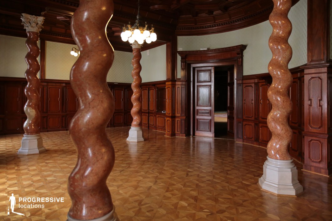 This distinctive hall features striking twisted marble columns that create a visually dynamic architectural statement. The deep wooden paneling along the walls and ceiling, combined with the polished parquet flooring, adds warmth and refinement to the space. Elegant lighting fixtures hang from the coffered ceiling, casting a soft glow that enhances the room’s intricate details. The symmetrical layout and open floor plan create a sense of grandeur, while the tall wooden doorway leads into an adjacent chamber, adding depth and continuity. This location is ideal for filming historical dramas, aristocratic meetings, or scenes set in a secretive, opulent environment with a unique architectural flair.