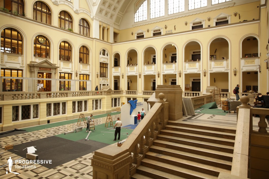 This expansive neoclassical atrium features soaring arched windows and balconies, creating an elegant, open atmosphere. The cream-colored walls are adorned with classical moldings, while the symmetrical design emphasizes balance and refinement. A central staircase with carved stone balustrades leads to the main floor, which showcases an intricate black-and-white geometric tile pattern. Large upper windows flood the space with natural light, highlighting the stately proportions. This location is ideal for filming academic settings, formal events, or institutional power dynamics, making it perfect for university dramas, historical reconstructions, or sophisticated corporate scenes.