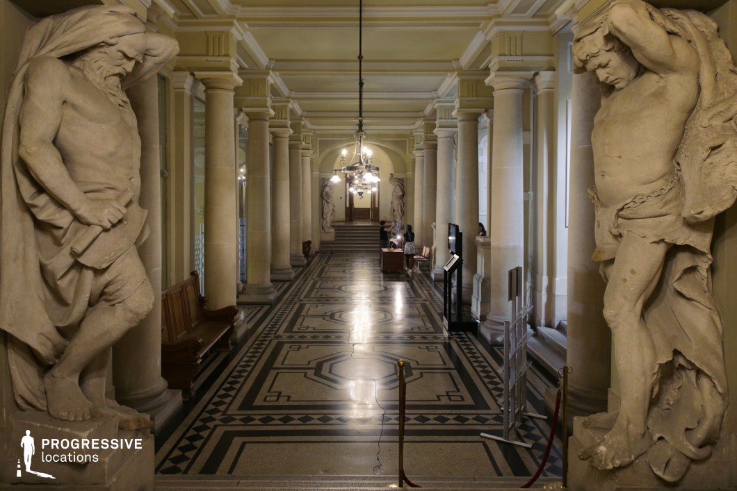 This stately neoclassical corridor features a symmetrical colonnade, high vaulted ceilings, and intricate black-and-white patterned flooring. Massive stone statues depicting mythological or allegorical figures frame the entrance, adding a monumental feel to the space. The polished marble-like surfaces and soft, ambient lighting enhance the refined atmosphere. Elegant chandeliers hang from the ceiling, casting a glow that highlights the architectural details. Wooden benches and decorative elements along the walls reinforce the historical significance of the setting. This location is ideal for filming period dramas, political intrigue, or scenes set in a grand courthouse, palace, or historic institution.
