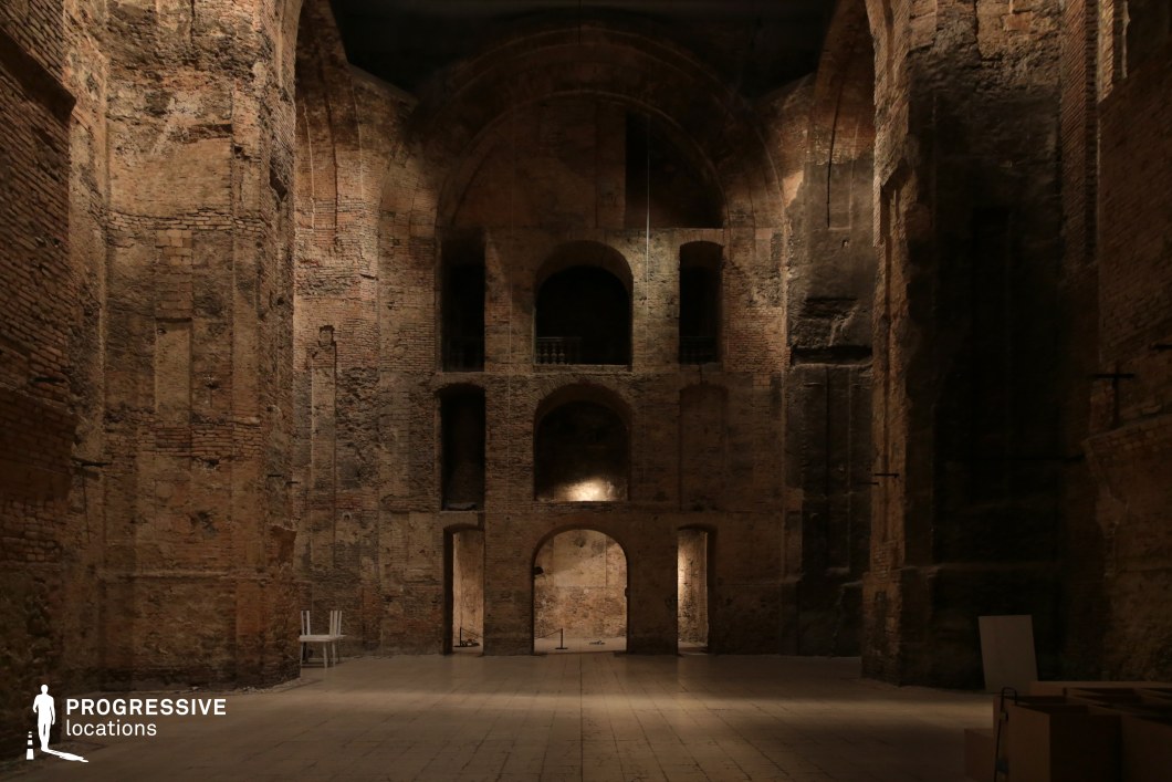 This vast, dimly lit space exudes a sense of mystery and historical depth, with towering weathered brick walls and imposing arched openings that suggest a once-grand structure now in a state of elegant decay. The uneven textures of the exposed masonry, combined with soft, focused lighting, create a dramatic interplay of shadows and depth. The high, vaulted ceiling and recessed alcoves enhance the monumental scale of the room, evoking the remnants of an ancient temple, fortress, or monastery. This location is ideal for filming historical dramas, fantasy epics, or atmospheric thrillers, offering a setting rich in mystery, isolation, and timeless grandeur.