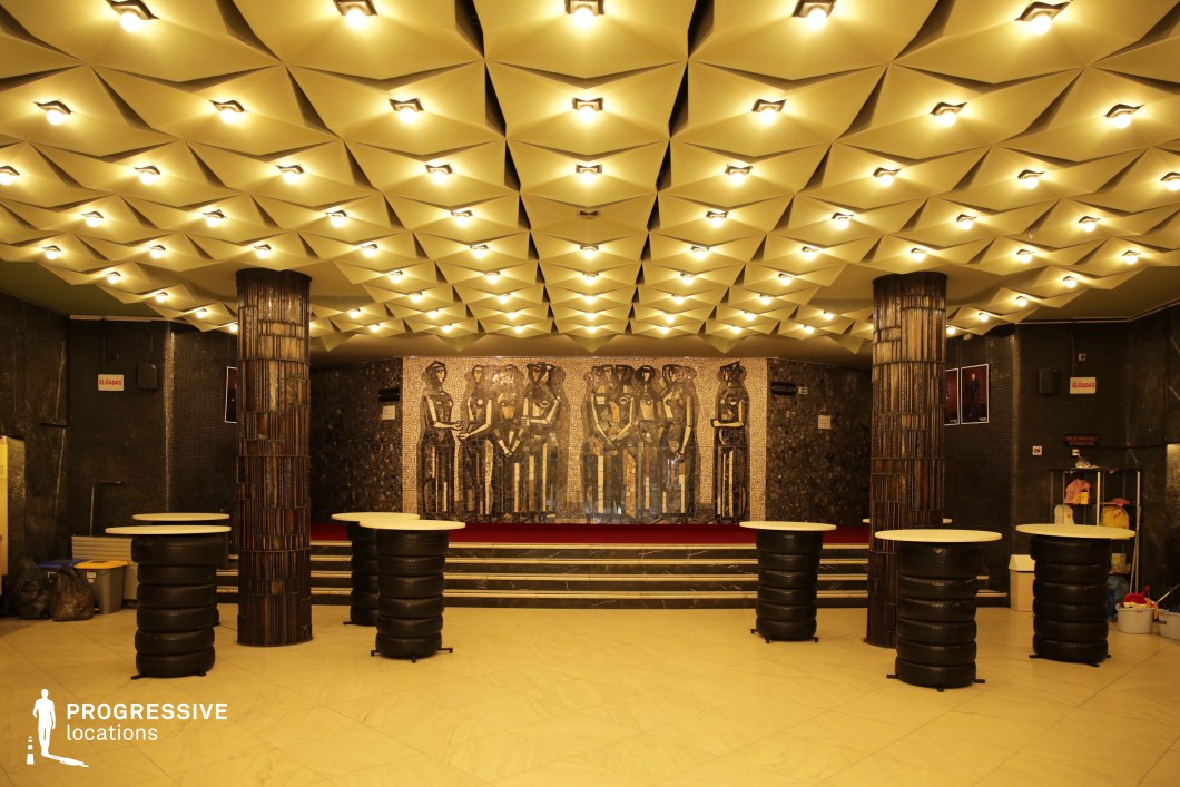This striking theater lobby features a geometric, three-dimensional ceiling with recessed lighting, creating a futuristic yet vintage aesthetic. The walls are adorned with dark mosaic tiles, with a focal point being a large metallic fresco depicting stylized human figures, adding an element of socialist realism or mid-century modern design. Two cylindrical columns with reflective surfaces anchor the space, while unconventional high-top tables with stacked tire-like bases contribute to the industrial-retro atmosphere. The grand staircase leading to the mural enhances the sense of depth and grandeur. This unique location is ideal for filming dystopian narratives, neo-noir thrillers, or retro-futuristic dramas, as well as for scenes requiring a striking, avant-garde backdrop.