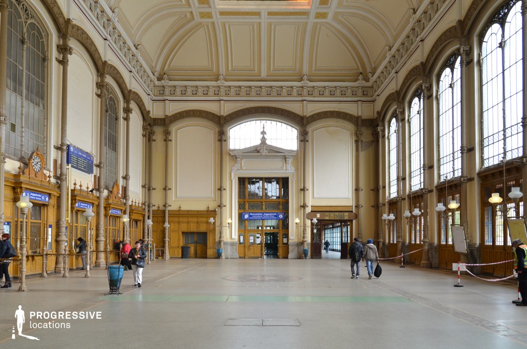This grand train station hall features soaring vaulted ceilings with intricate moldings, tall arched windows framed by decorative ironwork, and wooden ticket booths adorned with classical detailing. The symmetrical design is emphasized by large entryways, ornate columns, and elegant hanging lamps that contribute to a refined yet functional aesthetic. The warm tones of the wooden elements contrast with the neutral-colored walls, creating an inviting yet formal ambiance. This location is ideal for period films, travel-themed dramas, and suspenseful departures or reunions, offering a setting rich in architectural grandeur and cinematic depth.