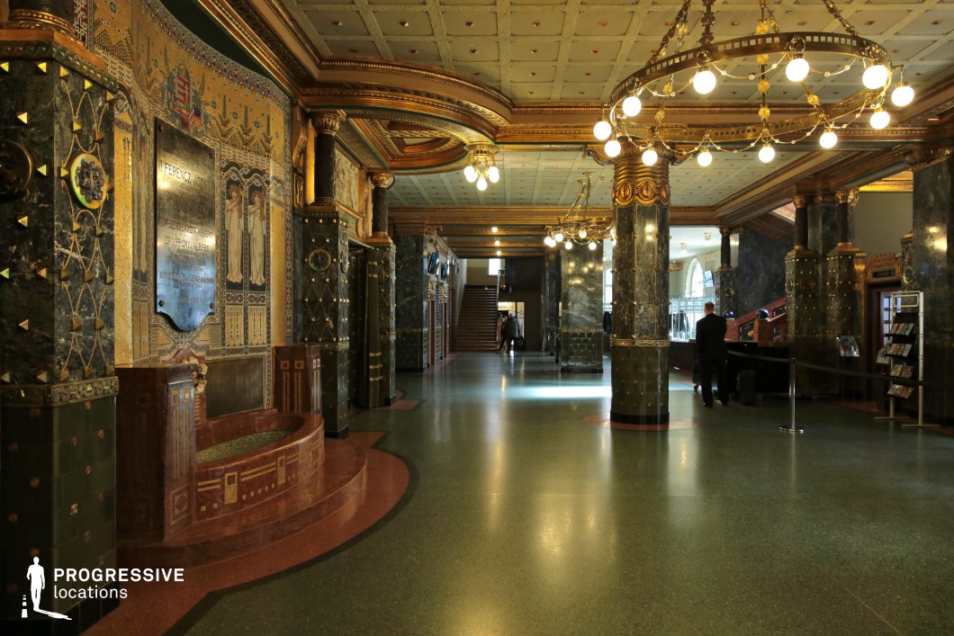 This grand Art Nouveau hall exudes an opulent yet sophisticated charm, featuring intricate mosaic detailing, polished dark green marble columns, and decorative gold accents. The ceiling, adorned with geometric paneling and suspended circular chandeliers, casts a warm, atmospheric glow over the space. The curved architectural elements, along with delicate tilework and stylized floral motifs, emphasize the fluidity characteristic of the Art Nouveau style. A striking mosaic wall panel with historical inscriptions and figurative artwork serves as a focal point, adding cultural depth. The hall’s open layout and sweeping staircase in the background create a sense of grandeur, making it ideal for filming historical dramas, period thrillers, or elegant high-society settings that require a richly detailed, visually engaging backdrop.