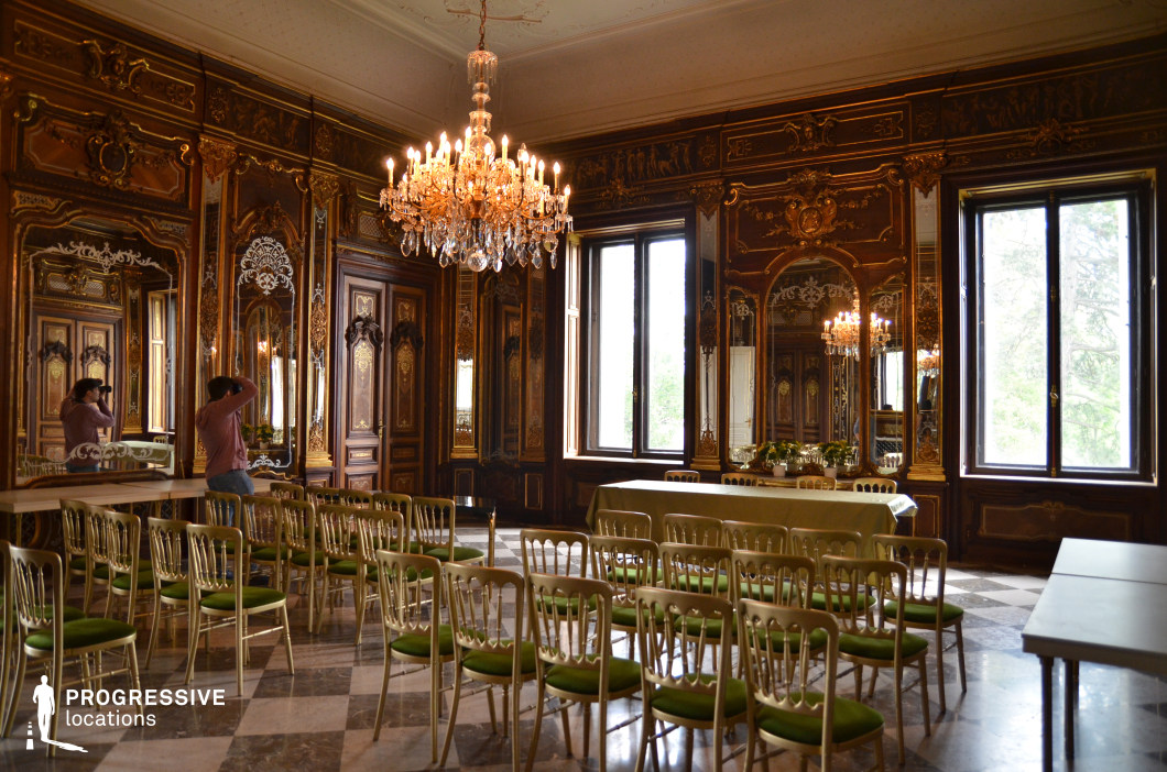 This grand hall features dark wood paneling with intricate gilded moldings, large mirrors framed in elaborate carvings, and a striking crystal chandelier that casts a warm glow over the space. The polished marble floor with a checkerboard pattern adds a refined contrast to the opulent decor. Tall windows allow natural light to filter in, illuminating the luxurious furnishings and gold-accented details. Rows of elegant chairs with green upholstery are arranged for a formal event, reinforcing the hall’s aristocratic ambiance. This location is ideal for filming historical dramas, royal ceremonies, or high-society gatherings, providing a sophisticated and regal setting.