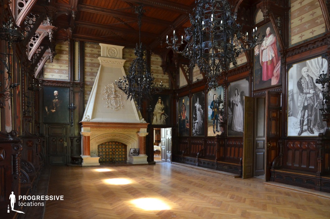 This striking hall exudes a Gothic Revival aesthetic, featuring dark carved wood paneling, elaborate wrought-iron chandeliers, and a grand stone fireplace adorned with intricate relief detailing. The high wooden ceiling, supported by decorative beams, enhances the dramatic atmosphere. Large aristocratic portraits line the walls, framed by rich, dark wood moldings, contributing to the historic grandeur. The polished parquet floor reflects the filtered natural light, adding warmth to the space. This location is ideal for filming historical dramas, gothic mystery scenes, or period-piece storytelling set in a noble residence, aristocratic estate, or secretive council chamber.