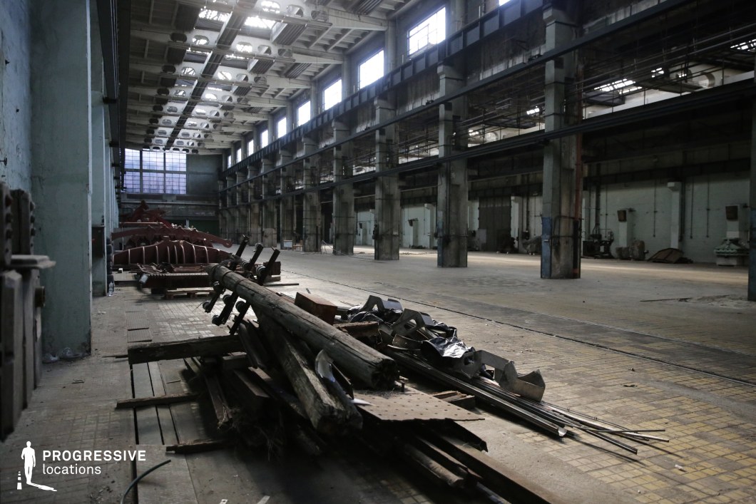 This expansive, abandoned warehouse features towering concrete pillars and a high ceiling with metal framework, giving it a raw and industrial aesthetic. Large, grime-covered windows allow limited light to filter through, casting shadows across the worn tiled and concrete flooring. The walls show signs of decay, with peeling paint and exposed wiring, while scattered debris and discarded metal structures hint at the space’s former use as a manufacturing or storage facility. The vast open layout and gritty atmosphere make this location ideal for filming action, dystopian, or post-apocalyptic scenes, as well as crime or thriller sequences requiring a desolate, tension-filled backdrop.