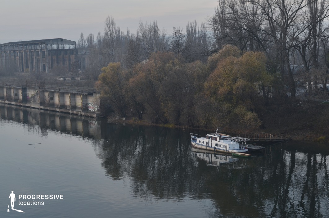 This riverside location features a decaying concrete embankment with graffiti-marked walls, an old industrial ruin in the background, and a cluster of leafless trees adding to the desolate atmosphere. A small, weathered boat is moored to a simple wooden dock, its reflection visible in the still water. The muted colors and abandoned structures contribute to a melancholic and eerie aesthetic, making this location ideal for filming psychological dramas, crime thrillers, or post-apocalyptic settings. The juxtaposition of industrial decay and natural overgrowth adds visual depth, enhancing the cinematic potential for atmospheric and suspenseful storytelling.