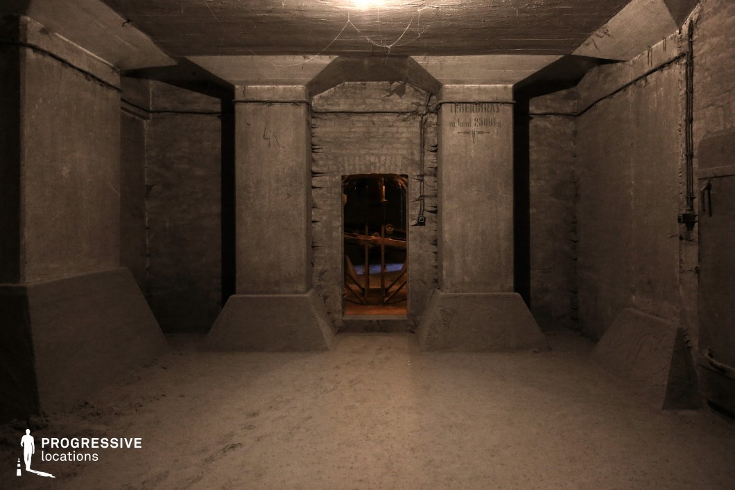 This dimly lit, industrial basement features thick, reinforced concrete columns and angular architectural supports, creating a bunker-like atmosphere. The rough-textured walls and ceiling are covered in dust and cobwebs, adding to the sense of abandonment. A small doorway framed by massive concrete beams leads to another chamber, where faint industrial machinery and warm, eerie lighting contrast with the cold, gray tones of the foreground. Exposed wiring and faded markings on the walls suggest a past functional purpose. This location is ideal for filming suspenseful, dystopian, or horror scenes, offering a confined, oppressive space perfect for mystery, captivity, or secret facility storylines.