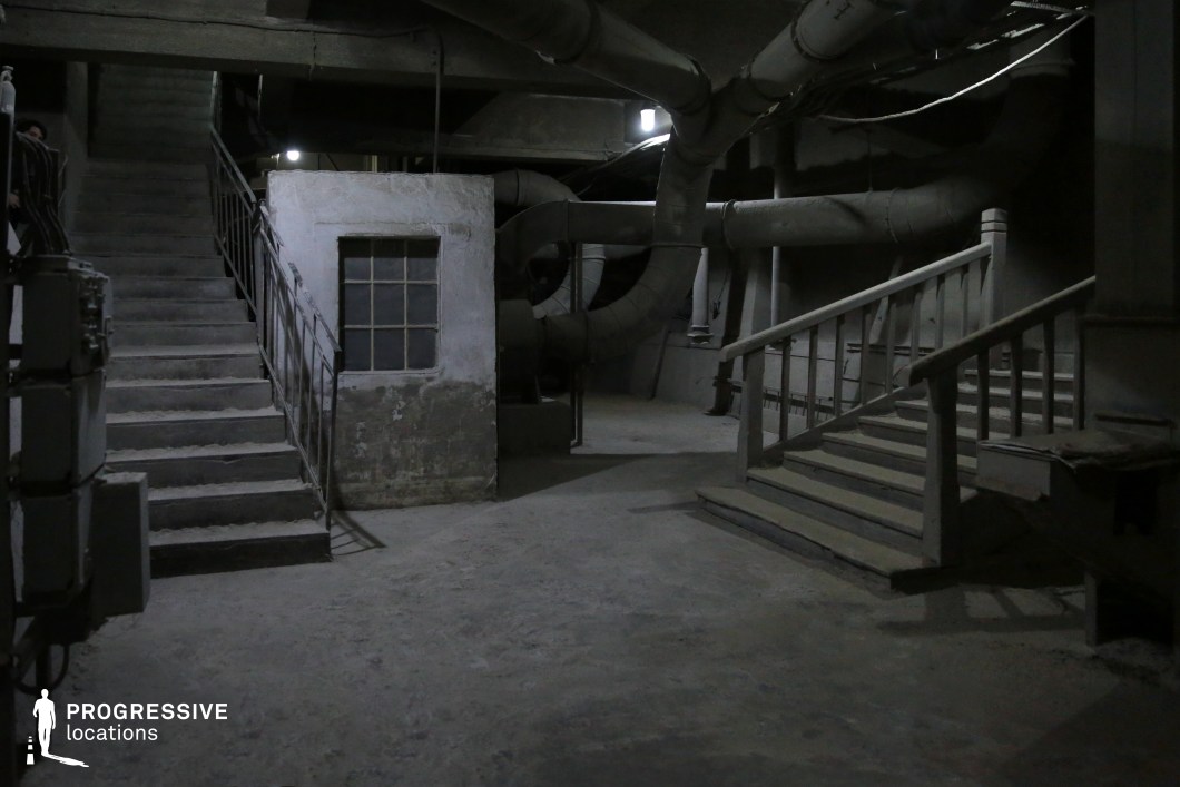 This eerie industrial basement features a labyrinth of exposed ventilation ducts, crumbling concrete walls, and two distinct staircases leading in different directions, adding a sense of mystery and depth. The dim lighting, aged wooden and metal railings, and dust-covered surfaces enhance the abandoned and unsettling atmosphere. A small window set into a deteriorating wall suggests a hidden or enclosed space, while the shadows cast by the overhead pipes create an ominous feel. This location is ideal for horror, thriller, or dystopian films, particularly for escape sequences, underground hideouts, or suspenseful confrontations.