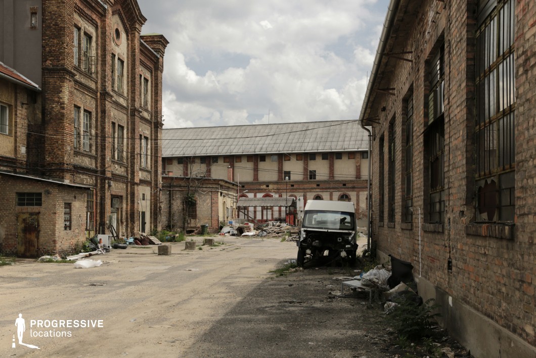 This desolate industrial courtyard is surrounded by aged brick buildings, featuring arched windows, boarded-up openings, and crumbling facades. The uneven ground is littered with scattered debris, while rusted metal bars and broken glass add to the atmosphere of decay. A large, imposing structure in the background, with a weathered roof and faded brickwork, suggests a former factory or storage facility. The neglected condition and muted color palette create a sense of abandonment and urban ruin. This location is ideal for filming post-apocalyptic, crime, or historical drama scenes, offering a raw, gritty setting for tense or atmospheric storytelling.