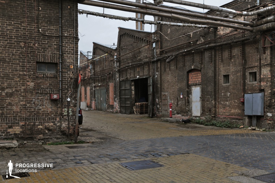 This atmospheric industrial alley is lined with weathered brick warehouses, their facades marked by peeling paint, exposed pipes, and rusted metal doors. Overhead, a network of corroded pipes stretches across the passage, reinforcing the utilitarian character of the space. The uneven cobblestone pavement, worn and patched in places, adds to the aged and gritty aesthetic. The sense of decay and neglect makes this location ideal for crime dramas, neo-noir, or dystopian productions, serving as a smuggling route, a hidden industrial district, or the setting for an underground black-market operation.