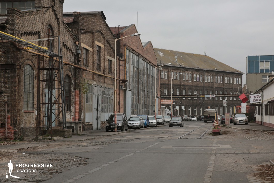 A weathered industrial street lined with old brick warehouses and factory buildings, featuring metal-framed windows, rusted pipes, and large steel doors. The road is worn with visible cracks and embedded tram tracks, reinforcing the location’s utilitarian past. In the background, a larger industrial complex with a yellowed rooftop and modern glass structures contrast with the aged facades. Overhead, exposed wiring and streetlights add to the gritty, urban feel. This location is ideal for film projects requiring an industrial, historical, or post-industrial setting, suitable for crime dramas, dystopian backdrops, or urban transformation narratives.