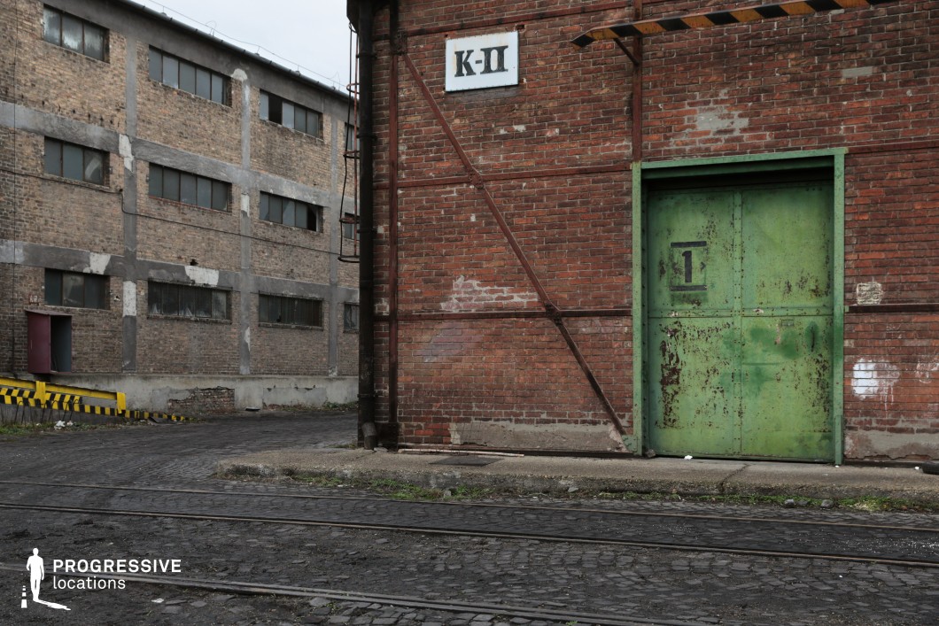 This weathered industrial site features a red brick warehouse facade reinforced with rusted metal braces, centered around a large, aged green metal door with peeling paint. A white sign with black lettering adds a functional, utilitarian feel. The adjacent structure, made of exposed brick and concrete, has long, narrow windows covered with grime, evoking an atmosphere of decay and abandonment. The uneven cobblestone ground, embedded with old rail tracks, suggests a former transport hub or factory loading area. The mix of rugged textures and muted tones makes this an ideal location for filming gritty crime dramas, post-apocalyptic settings, or historical industrial scenes, providing a raw, authentic backdrop for tense and atmospheric storytelling.