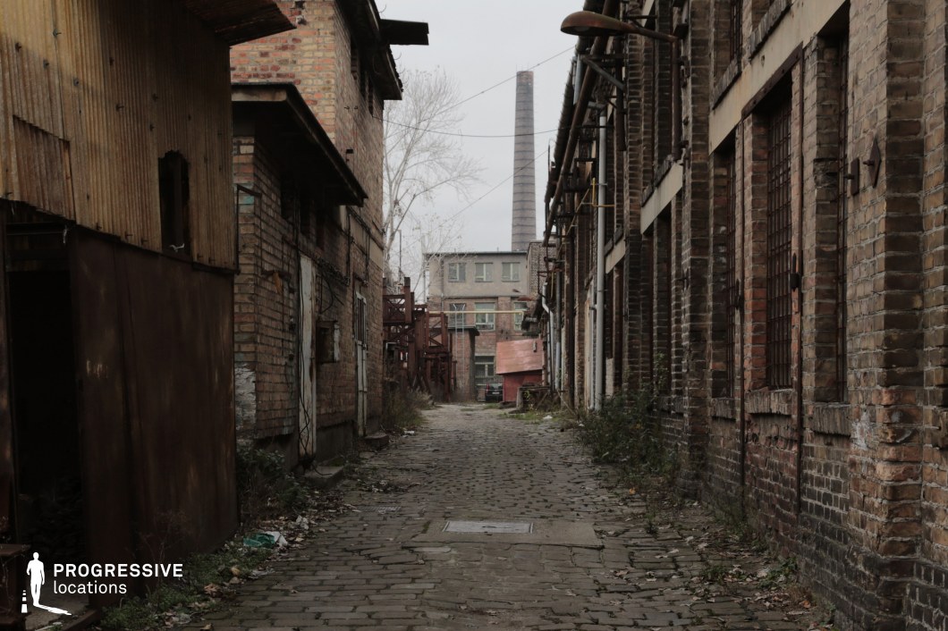 This desolate alleyway is framed by crumbling brick buildings, with rusted metal fixtures, broken windows, and overgrown vegetation creeping through cracks in the cobblestone path. Pipes and exposed wiring run along the walls, adding to the gritty, industrial decay. In the background, a towering smokestack rises above a distant factory structure, reinforcing the location’s abandoned, post-industrial atmosphere. The muted colors, rough textures, and sense of neglect make this an ideal filming location for crime dramas, dystopian thrillers, or post-apocalyptic settings, offering a raw, immersive environment perfect for high-tension or suspenseful scenes.
