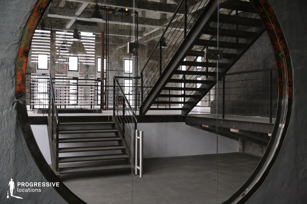 This industrial-style interior features a striking circular window framing a black metal staircase that leads to an open loft space. The exposed concrete walls, large grid windows, and hanging industrial lamps contribute to the raw yet modern aesthetic. The metal railings and open stair structure create a sense of depth, while the interplay of natural and artificial light enhances the contrast between the rugged textures and sleek, polished elements. The unique circular opening adds a cinematic perspective, making this location ideal for filming contemporary dramas, fashion shoots, or futuristic thrillers requiring a blend of industrial heritage and modern minimalism.
