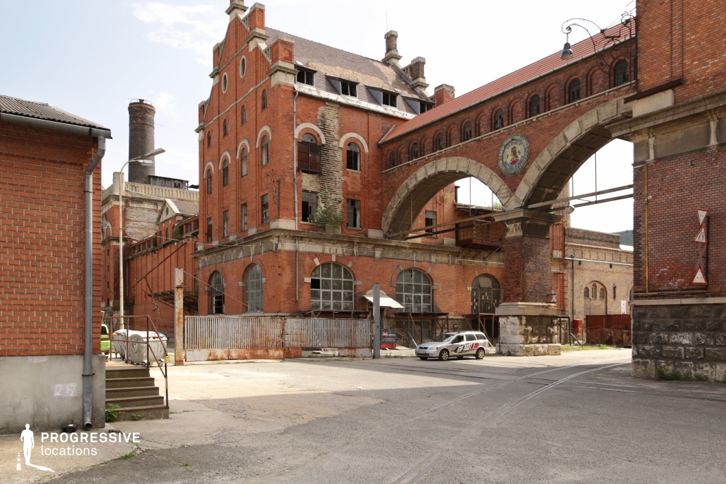 This grand industrial complex features red brick buildings with ornate detailing, large arched windows, and a striking elevated walkway connecting two structures. The aged facade, with exposed stonework and broken windows, suggests long-term disuse, while rusted metal gates and overgrown elements add to the atmosphere of decay. The intricate masonry and architectural flourishes evoke a historic European industrial style, making this location ideal for period dramas, historical thrillers, or neo-noir films, serving as an abandoned brewery, a forgotten factory, or a turn-of-the-century manufacturing hub.