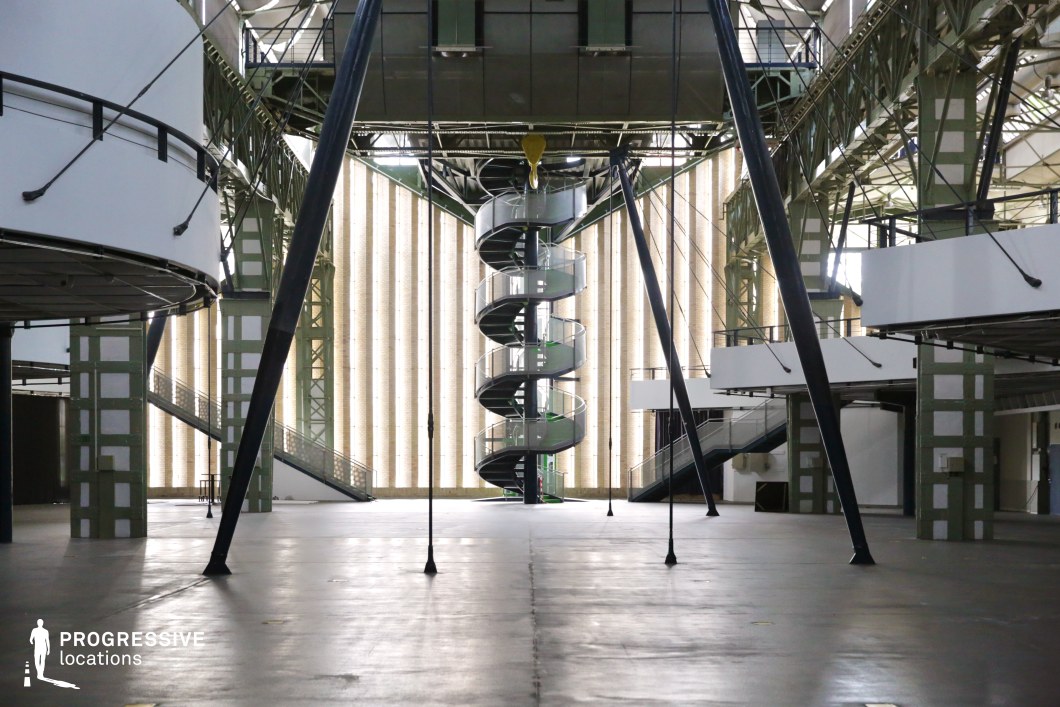 This vast industrial-style hall features a striking central spiral staircase encased in glass and metal, adding a futuristic and almost sci-fi aesthetic. The interior is defined by large steel beams, suspended walkways, and geometric structures, creating a sense of openness and precision. The walls are lined with tall, illuminated panels that diffuse light evenly, enhancing the modernist appeal. The polished concrete floor reflects the architectural details, further amplifying the high-tech, industrial atmosphere. This location is perfect for filming science fiction, cyberpunk, or high-stakes corporate thriller scenes, offering a sleek yet imposing backdrop for futuristic storytelling.