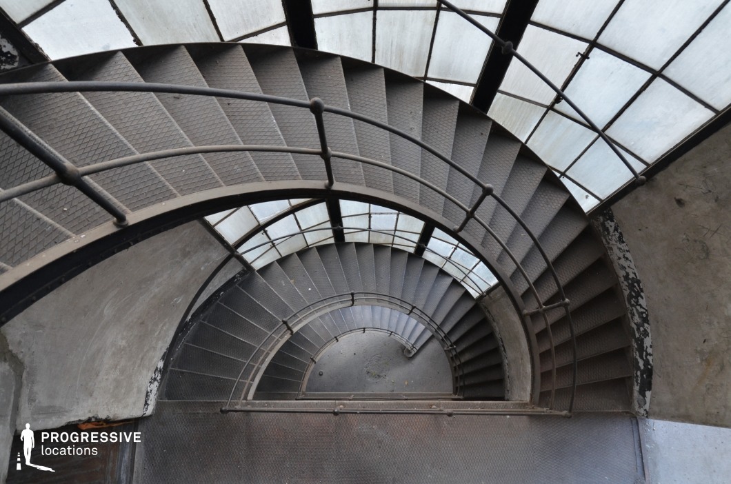 This striking industrial-style spiral staircase features dark metal steps with a textured surface and a curved railing, descending in a hypnotic geometric pattern. Encased within a partially translucent glass canopy supported by metal beams, the structure creates an interplay of light and shadow. The aged concrete walls show signs of wear, adding to the raw, utilitarian aesthetic. The confined yet dynamic space makes it an ideal location for suspenseful, psychological, or neo-noir film scenes, emphasizing mystery, pursuit, or dramatic character moments with visually engaging cinematography.
