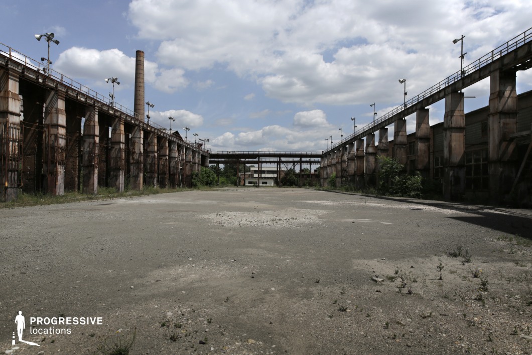 This expansive industrial site features two parallel rows of towering, rusted steel columns supporting an overhead walkway, creating a strong sense of symmetry and depth. The wide, cracked concrete ground is strewn with debris, adding to the abandoned, post-industrial feel. A distant smokestack and a small white building at the far end frame the vanishing point, enhancing the location’s desolate atmosphere. The combination of weathered metal, aged brick, and overgrown vegetation reinforces the site’s decay and former industrial significance. This setting is ideal for filming dystopian, post-apocalyptic, or crime thriller scenes, providing an eerie, raw backdrop for high-intensity or dramatic sequences.