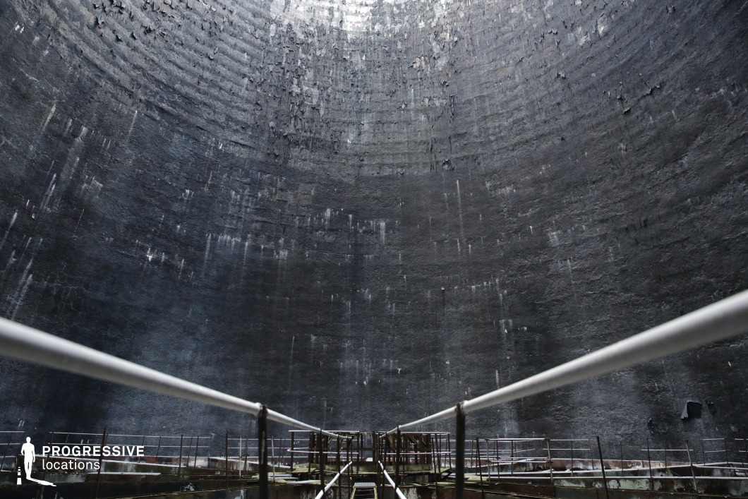 This vast, cylindrical industrial structure features towering concrete walls with a textured, weathered surface, marked by streaks of discoloration and peeling layers. A narrow metal walkway with railings extends toward the center, emphasizing the immense scale of the space. The curvature of the walls and the fading light at the top create a dramatic sense of enclosure, reinforcing an eerie, dystopian atmosphere. The industrial decay and sheer verticality make this location highly cinematic, ideal for sci-fi, post-apocalyptic, or thriller productions, serving as a secret research facility, an abandoned power plant core, or a surreal, otherworldly environment.